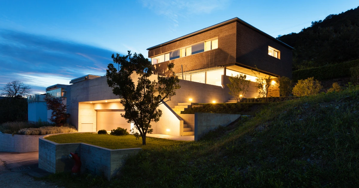 A contemporary house illuminated with warm lights at dusk, surrounded by greenery.